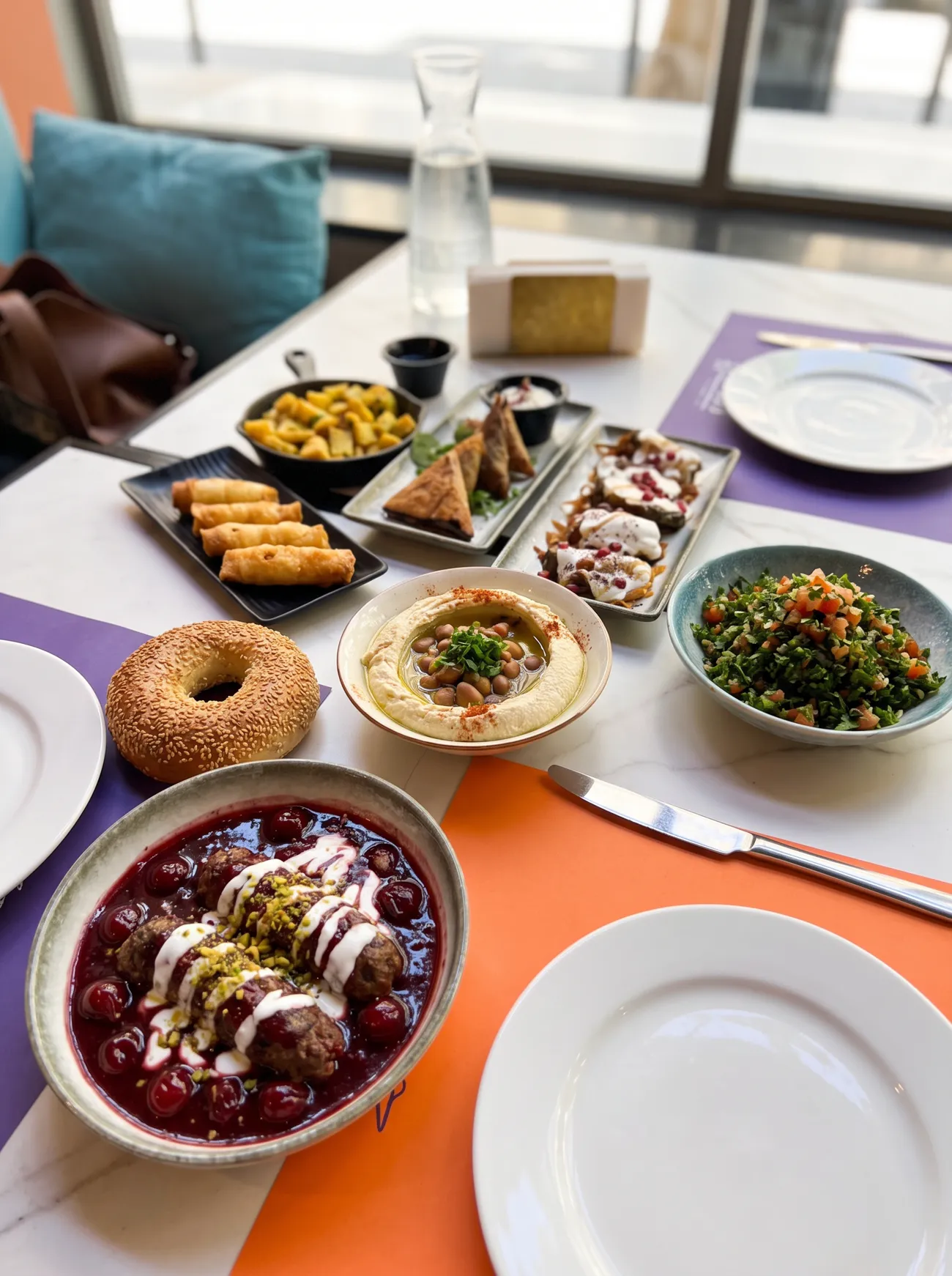 Kaftet karaz in cherry sauce, hommus with foul, tabbouleh, cheese rolls, batata harra, grape leaves with yogurt and a sesame kaak on orange and purple placemats